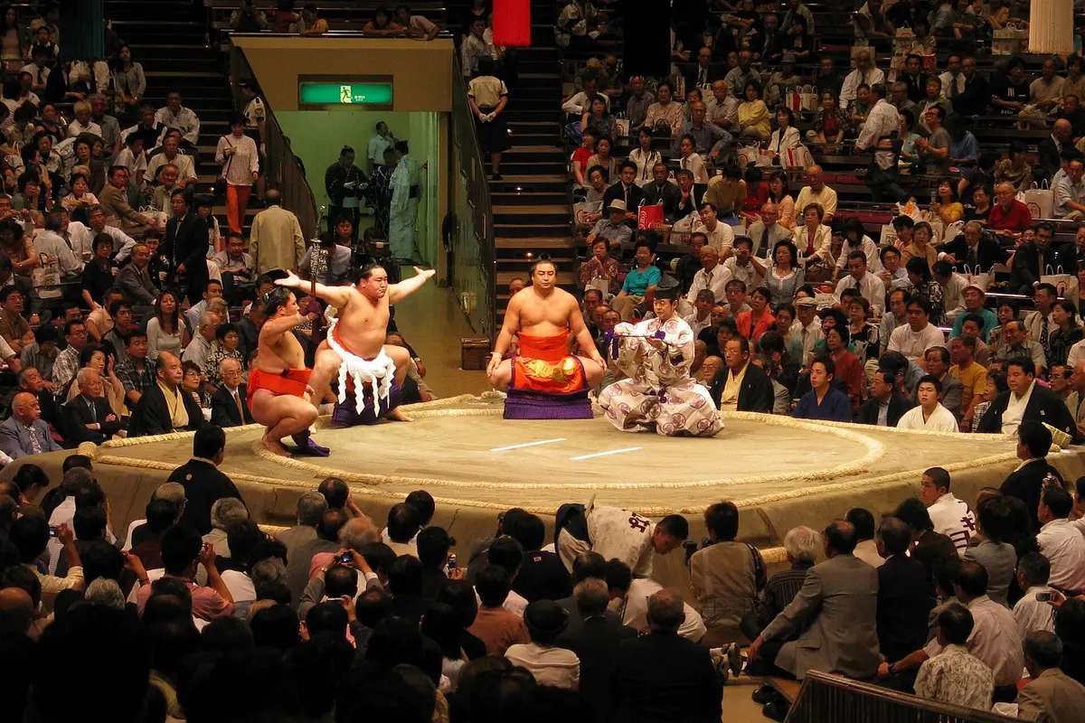 The raised clay ring at the Kokugikan with straw bales marking the circle and two white starting lines, a wrestler with arms outstretched and a kneeling referee beside him