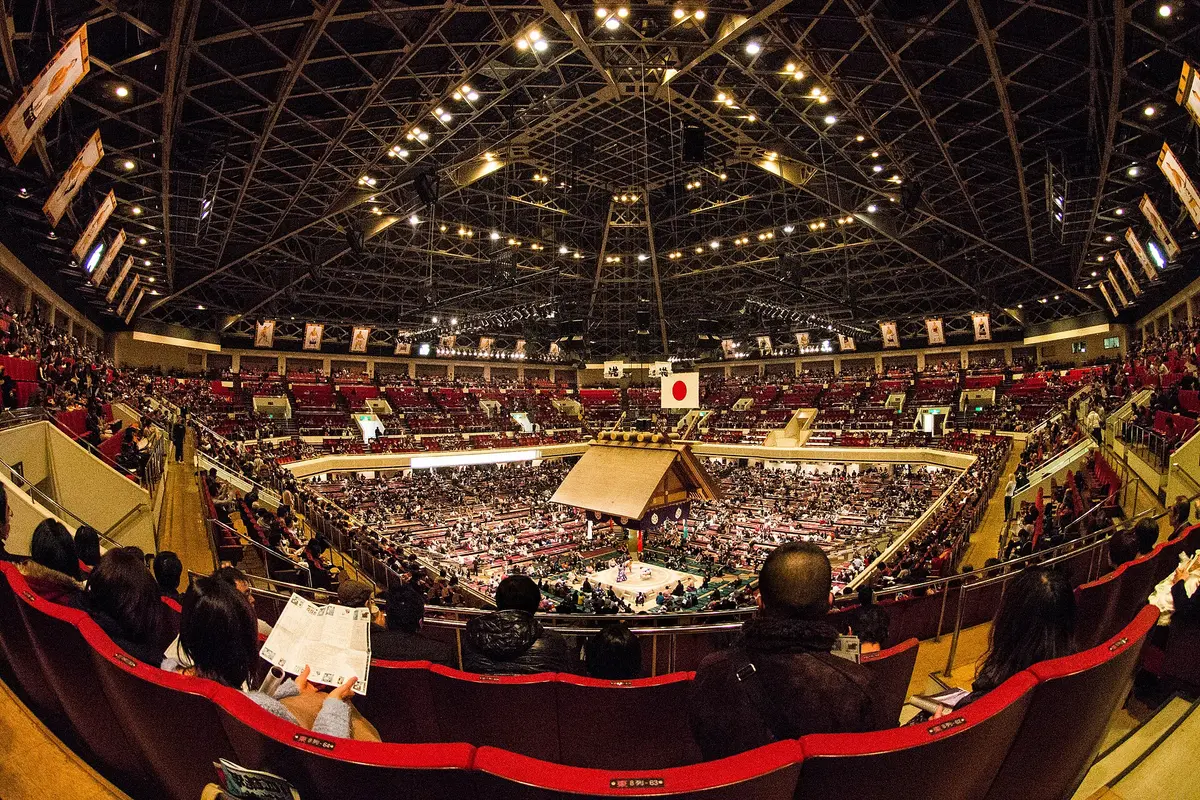 A wide view inside the Ryogoku Kokugikan with the shrine-style roof over the ring, floor boxes around it and red chair seats in the tiers above
