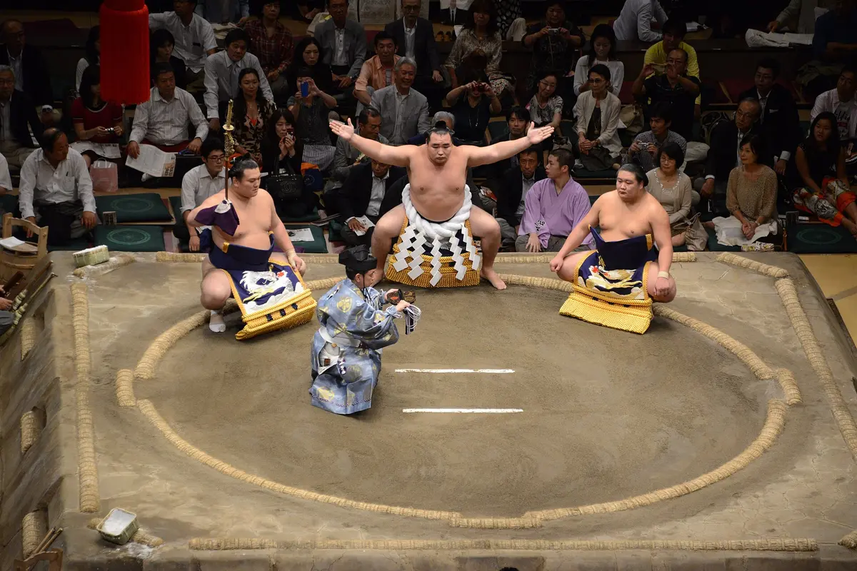 A yokozuna with arms stretched out performing the ring-entering ceremony in the clay ring, flanked by two attendants in embroidered aprons, with spectators behind